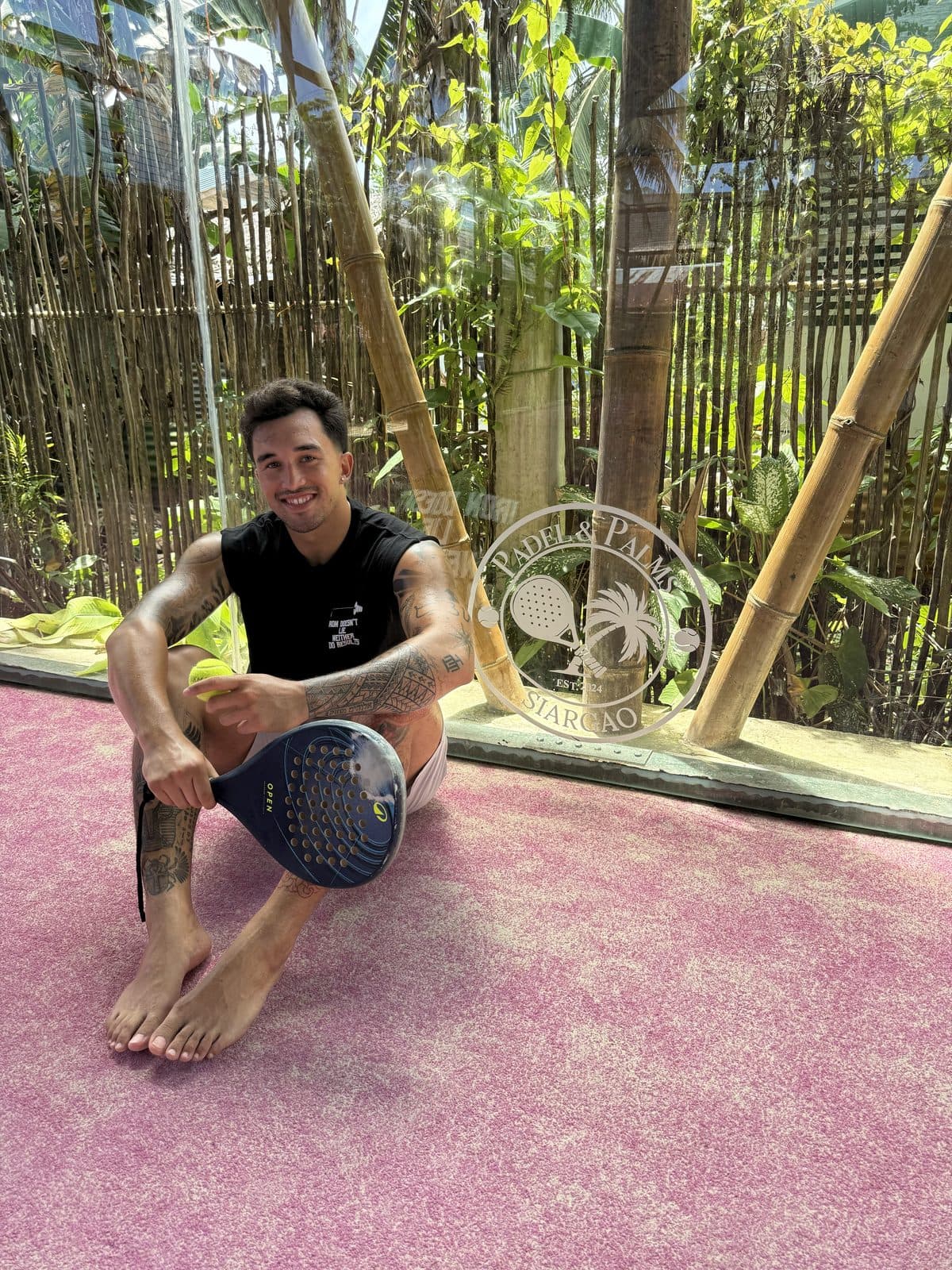 Ezekiel Contreras-Forrest sitting on a padel court in Siargao, Philippines, holding a racket and ball, with bamboo and jungle visible through the glass wall behind him